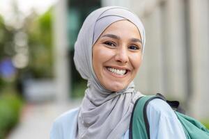 de cerca retrato de musulmán hembra estudiante en hiyab, mujer con mochila detrás su espalda sonriente y mirando a cámara, en pie fuera de Universidad edificio instalaciones. foto