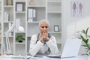 A female doctor wearing a hijab looks thoughtful and concerned while sitting at her desk in a modern clinic office, surrounded by medical books and a laptop. photo