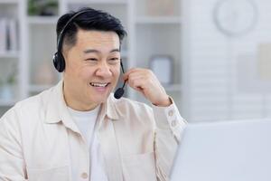 A cheerful Asian man wearing a headset smiles while working from his home office. He is interacting and providing customer support with positivity and professionalism. photo