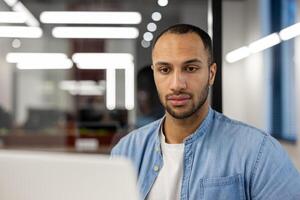 Close-up photo of a young hispanic man in a blue denim shirt, sitting at a desk in an office and looking seriously at a laptop screen.