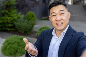 Close-up portrait of a young Asian man in a business suit standing outside an office center and talking on the camera of a phone, he is holding in his hands. photo