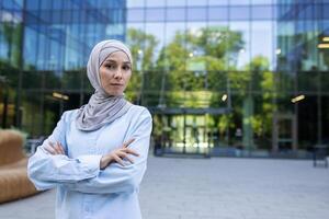 profesional musulmán mujer con brazos cruzado en frente de moderno corporativo arquitectura, exudando confianza y determinación. foto