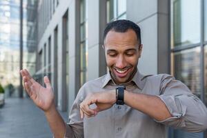 Young successful arab businessman talking on smart watch and smiling, app developer programmer walking outside modern office building, worker in casual shirt photo