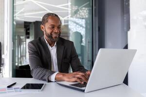 Successful mature african american businessman at work inside office, experienced boss working with laptop, man smiling satisfied with achievement results. photo