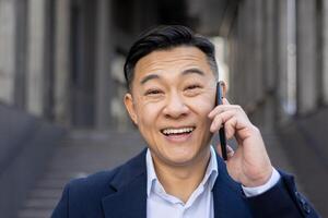 alegre asiático empresario por casualidad vestido en un traje haciendo un contento teléfono llamada mientras en pie al aire libre. foto