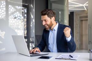 Happy young businessman sitting in the office at the table and looking at the laptop screen, rejoices at the success, received information, showing a victory gesture with his hand. photo