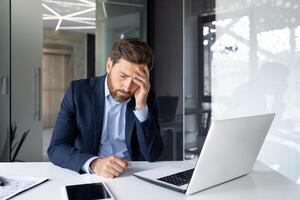 Businessman feeling stressed and overwhelmed at work, hand on forehead, sitting at desk with laptop in modern office. photo