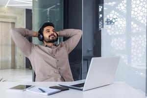 Indian young man relaxing at the workplace, satisfied with the finished project and the result. He sits relaxed at the table, wearing headphones, with his hands behind his head. photo