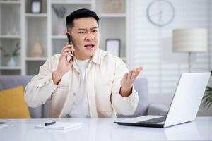 Angry angry and upset man talking on phone at home, asian working remotely from home office sitting at table in living room, freelance businessman at work. photo