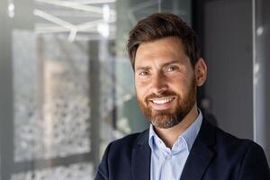 A professional portrait of a smiling businessman with a beard, standing in a well-lit, contemporary office environment. photo