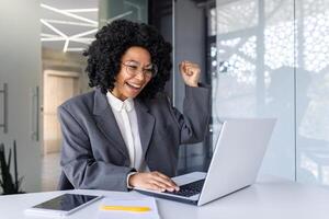 Successful african american woman at workplace inside office, business woman winner got good result and achievement at workplace, reading notification using laptop. photo