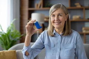 sonriente mujer con azul inhalador en mano mirando a cámara mientras sentado en Doméstico atmósfera. arrugado hembra consiguiendo usado a diario rutina para salud y bienestar con crónicamente enfermedad. foto