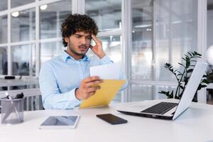 joven empresario a trabajo dentro oficina recibido correo sobre notificación mensaje con malo noticias, Hispano hombre triste leyendo letra documento sentado a mesa con ordenador portátil. foto