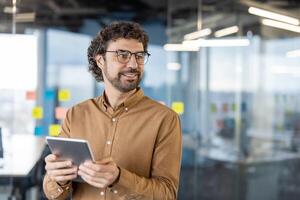 Confident male with curly hair looking aside with curious face while keeping electronic device in hands. Professional manager watching as colleagues passing by in hall of modern office. photo