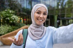 A cheerful student in a hijab takes a selfie, possibly on a call, amidst a vibrant cityscape. photo