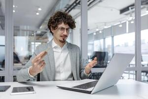 A young adult businessman is shown feeling perplexed at his workplace, a modern office, suggesting the challenges of problem-solving and decision-making that office workers often face. photo