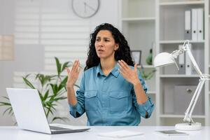 un incómodo mujer de negocios sensación también caliente en un oficina configuración, gesticulando en angustia cerca su espacio de trabajo foto