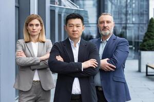 Successful and serious diverse team of three business people, man and woman focused looking at camera with arms crossed, portrait of co-workers outside office building photo