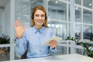 Positive office worker with headset waving palm in greeting gesture to camera and smiling friendly. Attractive female holding digital tablet and getting ready for providing customer support. photo