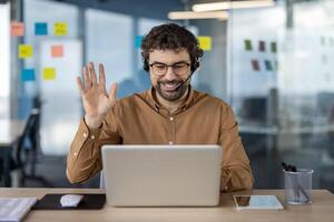 Professional man in a casual business setting using laptop for a cheerful conference with colleagues or clients, waving to the camera. photo