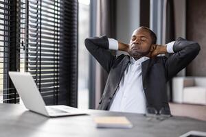 A professional middle-aged black businessman sits back in his office chair with his hands behind his head, eyes closed, embodying relaxation and stress relief amidst a busy work environment. photo