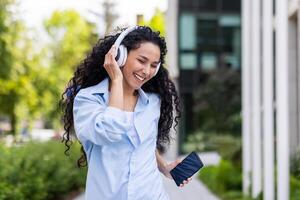 un alegre, pelo rizado mujer es atrapado en un momento de alegría mientras escuchando a música en su blanco auriculares, participación un teléfono inteligente, al aire libre con verdor y un edificio en el antecedentes. foto