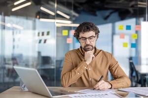 Serious thinking businessman behind paper work inside office, man fills forms, reports and invoices. An employee uses a laptop for paperwork. photo