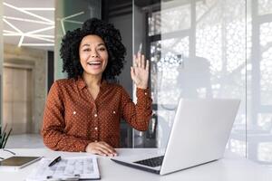 sonriente joven africano americano mujer trabajando en el oficina utilizando un computadora portátil, sentado a el escritorio, ondulación y saludo a el cámara. foto