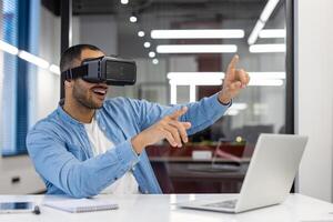 Smiling Indian man wearing virtual reality headset, interacting with digital content in a modern office environment. photo