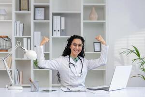 un alegre latino mujer médico con Rizado pelo levanta su brazos en un triunfante pose, celebrando un profesional éxito en su médico oficina. foto