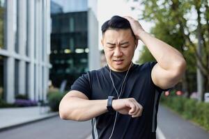 Upset young asian man standing in the middle of the street and looking at the smart watch, not happy with the result, holding his head in frustration. photo