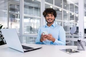 un joven latín americano empresario se sienta en el oficina a un escritorio y usos un móvil teléfono, escribe un mensaje, búsquedas para información en el Internet, cheques social redes sonriente a el cámara. foto