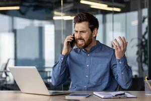 enojado y irritado joven empresario sentado en el oficina a el escritorio y hablando en el teléfono mientras ondulación su manos. foto