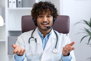 cámara web vista, joven médico con auriculares teléfono utilizando ordenador portátil para llamar, médico alegremente y simpático consultante pacientes, sonriente y mirando a cámara, trabajando dentro clínica médico habitación. foto