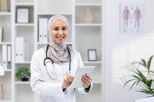 Beautiful arabic medical woman standing in workspace with digital tablet in hands. Confident female with stethoscope looking camera with smile and using gadget for communication with patients. photo