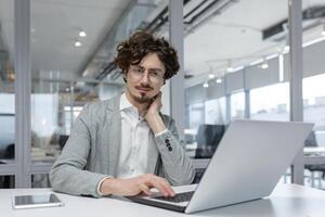 retrato de un contemplativo joven empresario con Rizado pelo trabajando diligentemente en su ordenador portátil en un moderno oficina ajuste. foto