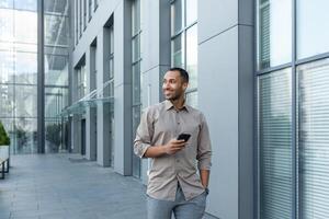 Smiling hispanic man walking down street near modern office building, freelancer businessman looking away holding mobile phone photo