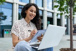 sonriente joven Hispano mujer trabajando en su ordenador portátil fuera de un oficina edificio, tomando notas durante un almuerzo descanso. foto