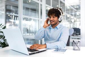 Young successful programmer inside modern office working with laptop, man in blue shirt smiling and happy, listening online music in headphones, audio books and podcasts, businessman satisfied work. photo