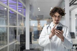 un médico en un blanco laboratorio Saco y estetoscopio usos un célula teléfono dentro un médico oficina. grave hombre leyendo social medios de comunicación foto