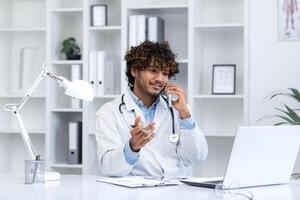 Young successful hispanic doctor inside the clinic in the medical office joyfully communicates talking on the phone, the clinic worker sits at the desk working with a laptop, consults patients. photo