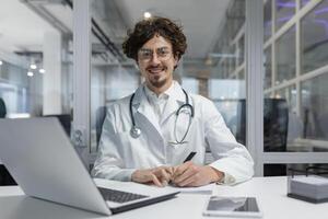 A doctor wearing a white lab coat and stethoscope is seated at a desk, using a laptop.Man smile happy writing report looking camera photo
