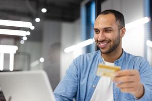 un joven adulto masculino participación un crédito tarjeta, pretendiendo a hacer un en línea compra mientras trabajando en su ordenador portátil en un contemporáneo oficina ajuste. foto