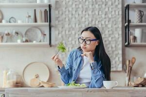 retrato de un joven hermosa asiático mujer sentado en el cocina a el mesa, participación un Fresco verde ensalada en su tenedor, suspirando, mirando a eso infelizmente cansado de haciendo dieta, odia vegetarianismo. foto