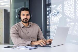 Portrait of a serious young Indian man businessman, freelancer working in the office with a laptop. Sitting at the table in headphones and looking at the camera. photo