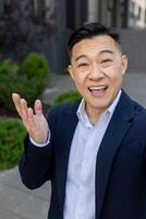 Happy Asian man standing outside in business suit and talking on call gesturing with hands. Vertical photo. photo