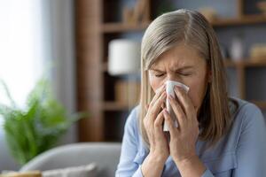A mature woman experiencing cold symptoms, sneezing into a tissue while sitting in a cozy living room. Health and wellness concept. photo