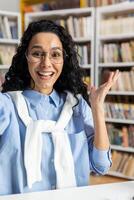 Joyful Hispanic Latino student wearing glasses in a library, smiling and gesturing with a sense of accomplishment and happiness. photo