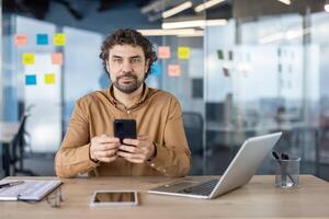 un enfocado de edad mediana profesional hombre usos un teléfono inteligente a su escritorio en un brillantemente iluminado oficina, mostrando productividad y conectividad. foto