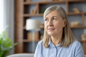 A serene mature woman with light hair sits comfortably in her well-decorated living room, gazing thoughtfully into the distance. photo